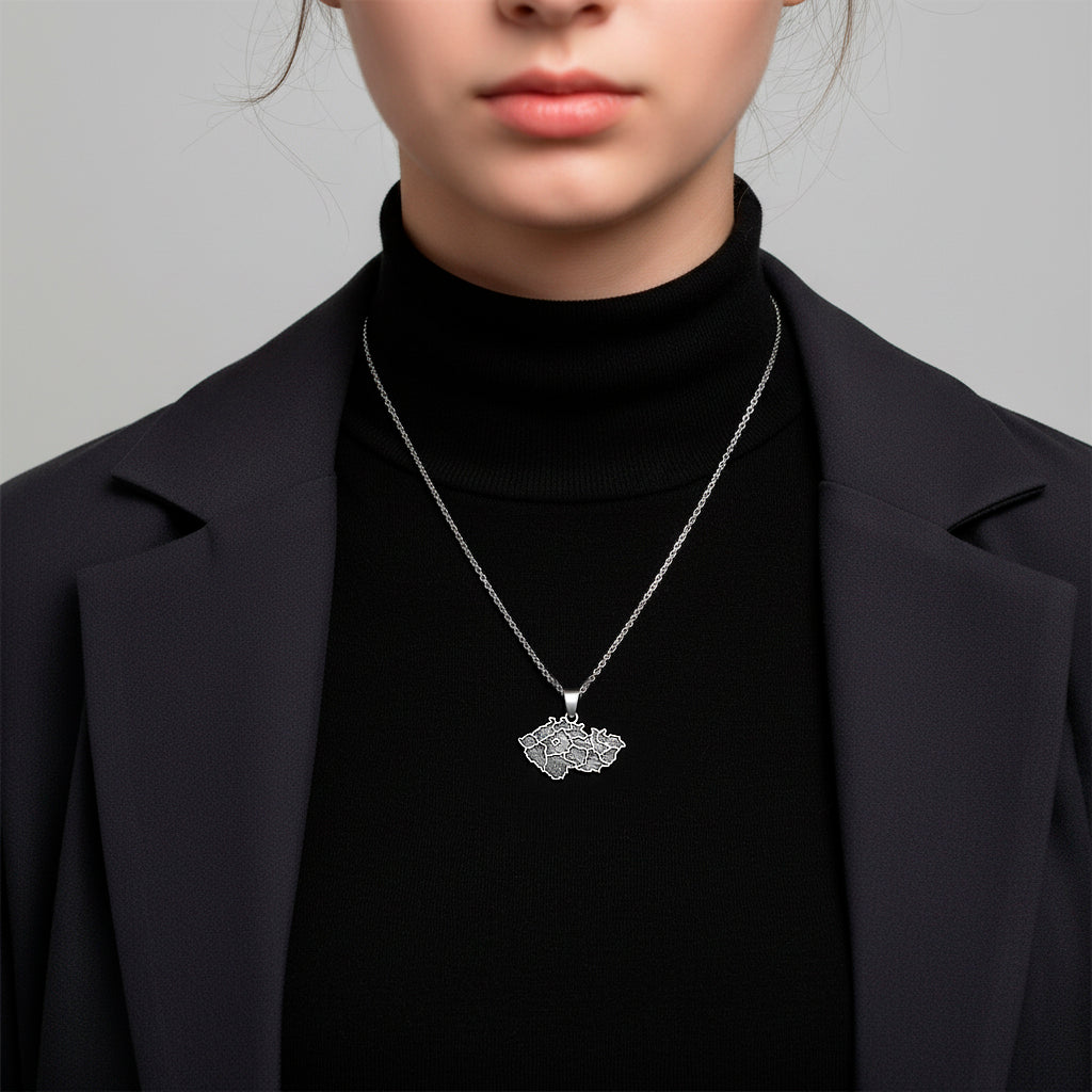 A handcrafted sterling silver pendant shaped like the map of the Czech Republic with the engraved phrase “Já❤️Česko.” The pendant measures 27 mm in width and 24 mm in height with the bail, weighs 3 g, and features a smooth polished edge with dark engraved details. It is displayed resting on a soft dark fabric cushion with gentle lighting that highlights the texture of the metal and the elegant contours of the design.