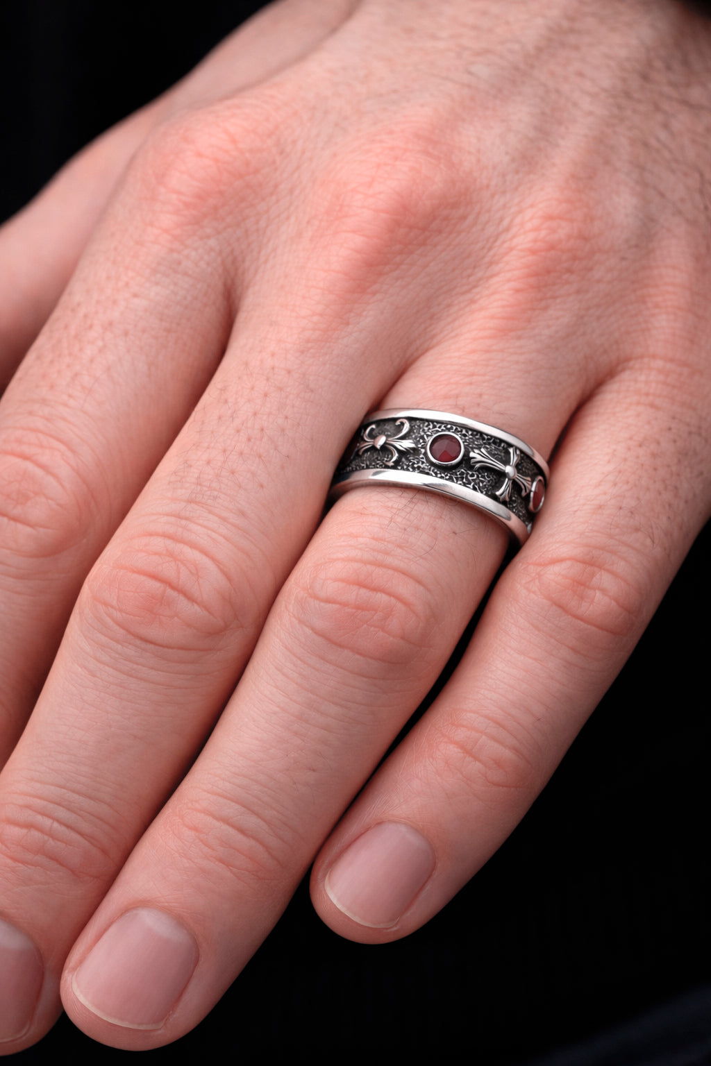 A close-up of a person's hand wearing a silver ring with a decorative design. Sterling silver Fleur De Lis ring with deep red garnet stones, featuring an oxidized textured finish and polished heraldic symbols. Medieval gothic band ring inspired by knightly and royal motifs. Solid 925 silver statement ring suitable as a wedding band or symbolic jewelry piece. Elegant, bold, and historically inspired design for men or women, perfect for gothic style, medieval fashion, and unique meaningful gifts.
