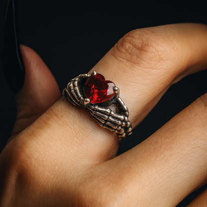 A close-up of a person's hand wearing a ring with a red gemstone and a skeletal hand design.