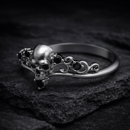 A silver ring with a skull design and black gemstones is displayed against a dark background.