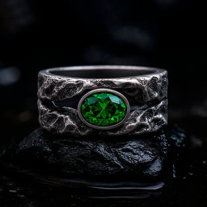 A silver ring with a large green gemstone, possibly a diamond, set in a textured metal band, placed on a black surface.