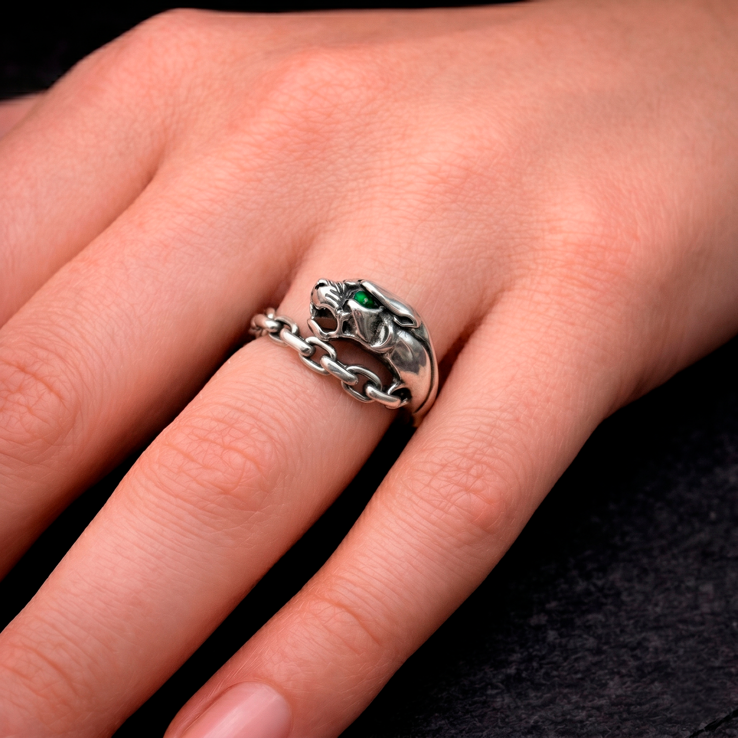 Highly detailed premium editorial photography of a sterling silver panther ring on a woman's finger. Handcrafted oxidized silver signet ring featuring a ferocious wild cat head with a vibrant green gemstone eye and an unclosed chain band. Punk gothic jewelry gift