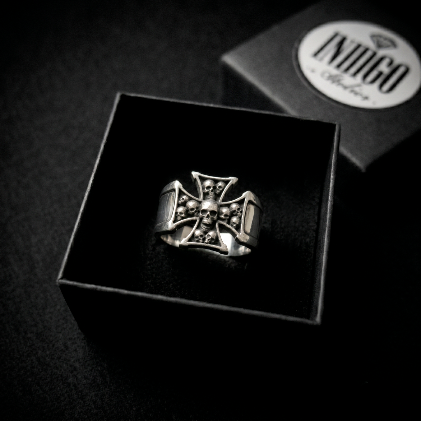 A silver ring with a skull and crossbones design is displayed in a black box, with a logo visible in the background.