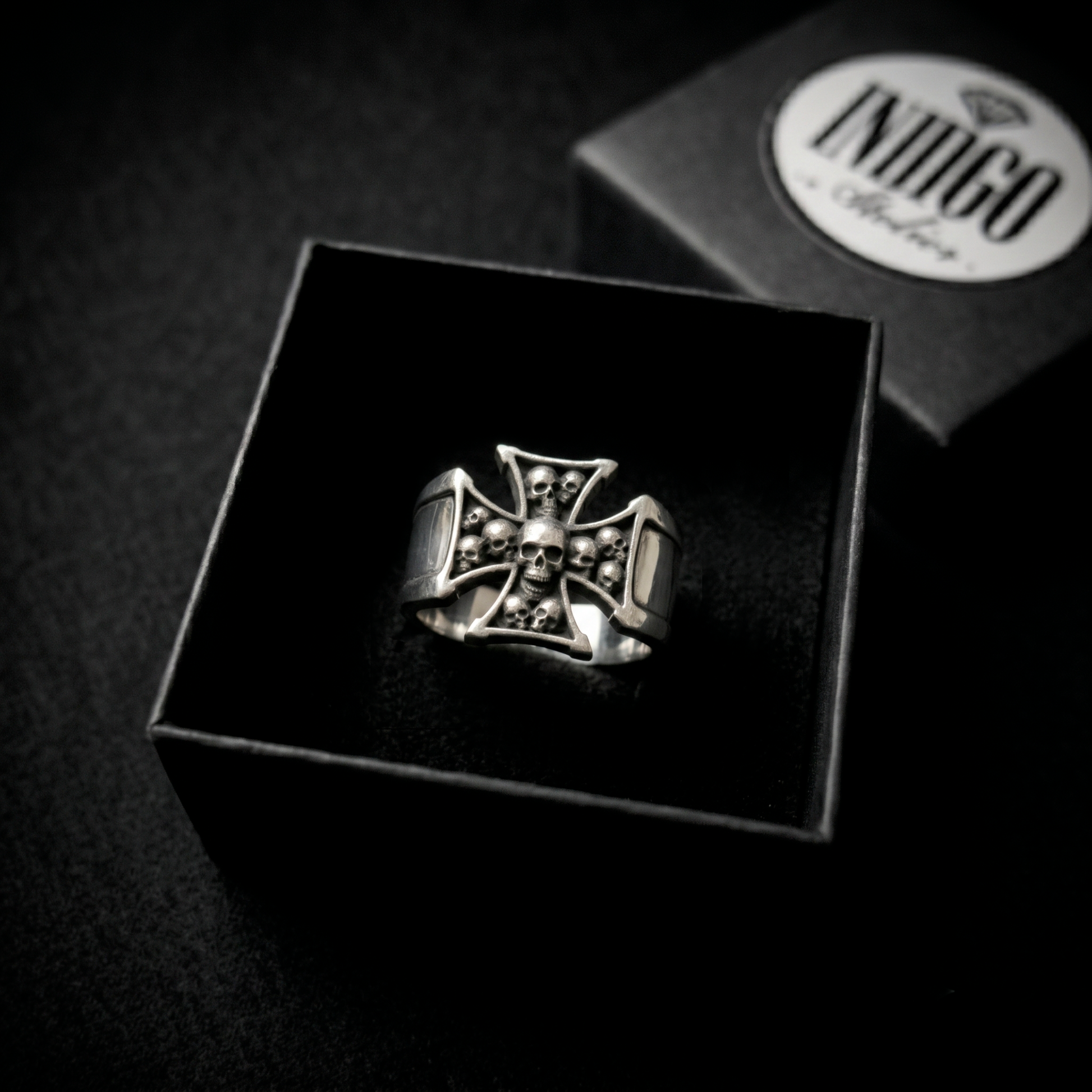 A silver ring with a skull and crossbones design is displayed in a black box, with a logo visible in the background.