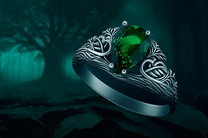 A silver ring with a large green gemstone, possibly an emerald, is displayed against a dark, moody background with a tree silhouette.