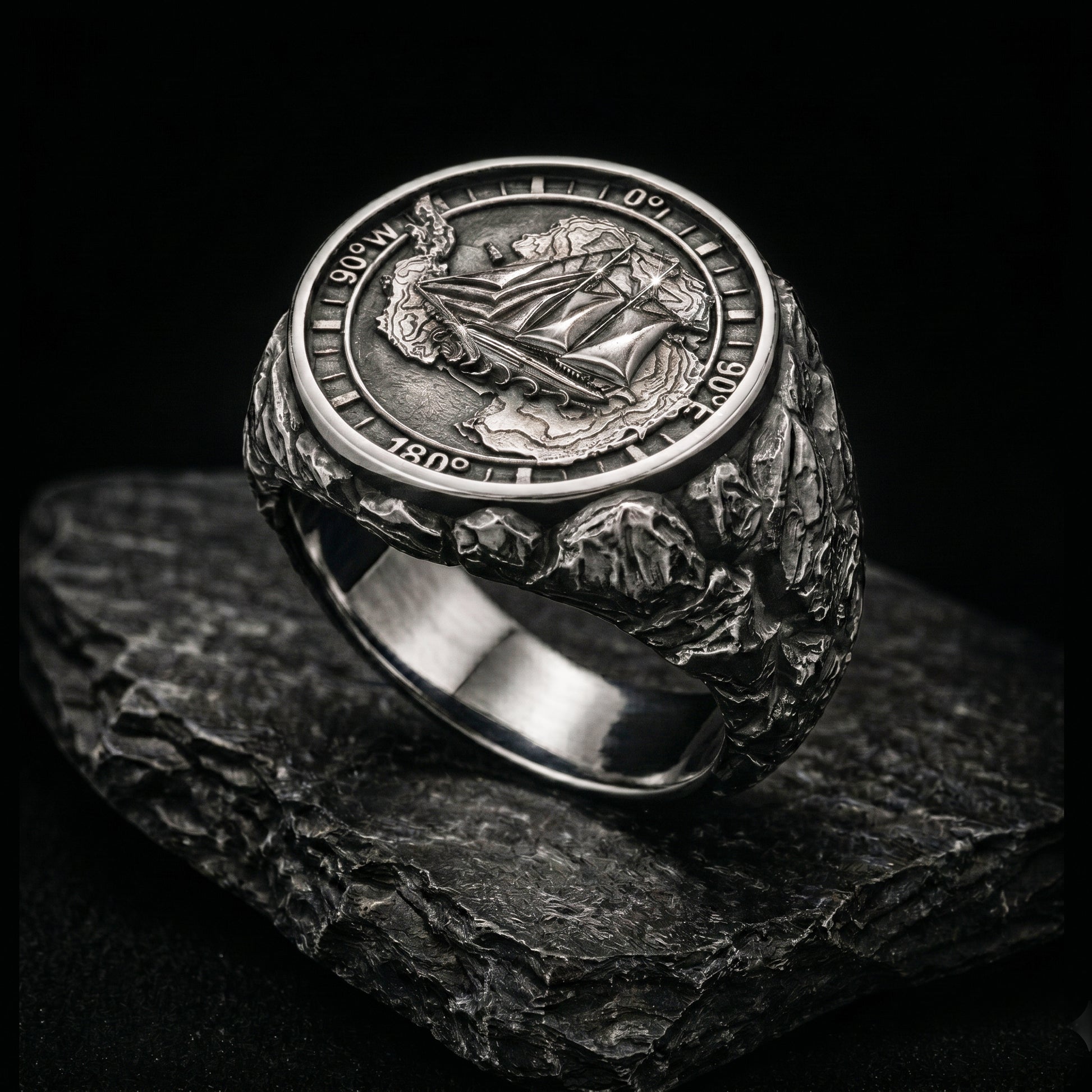 Antarctica explorer signet ring in solid sterling silver 925 featuring a detailed sailing ship over a map of Antarctica, surrounded by directional markings. The ring has a deep oxidized finish with strong contrast, highlighting fine handcrafted textures and relief details. Shot on a dark stone background to emphasize its bold, masculine design, adventure theme, and expedition spirit. Unique nautical jewelry piece inspired by travel, ocean journeys, and exploration beyond the known world.