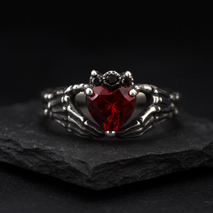 A ring with a red gemstone, resembling a heart, is displayed against a dark background.