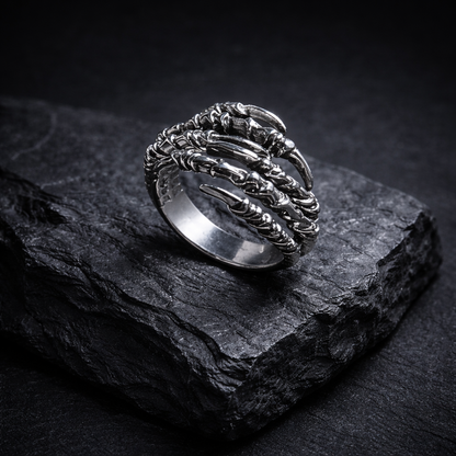 A silver ring with intricate designs rests on a dark, textured surface, such as a rock or a slate.