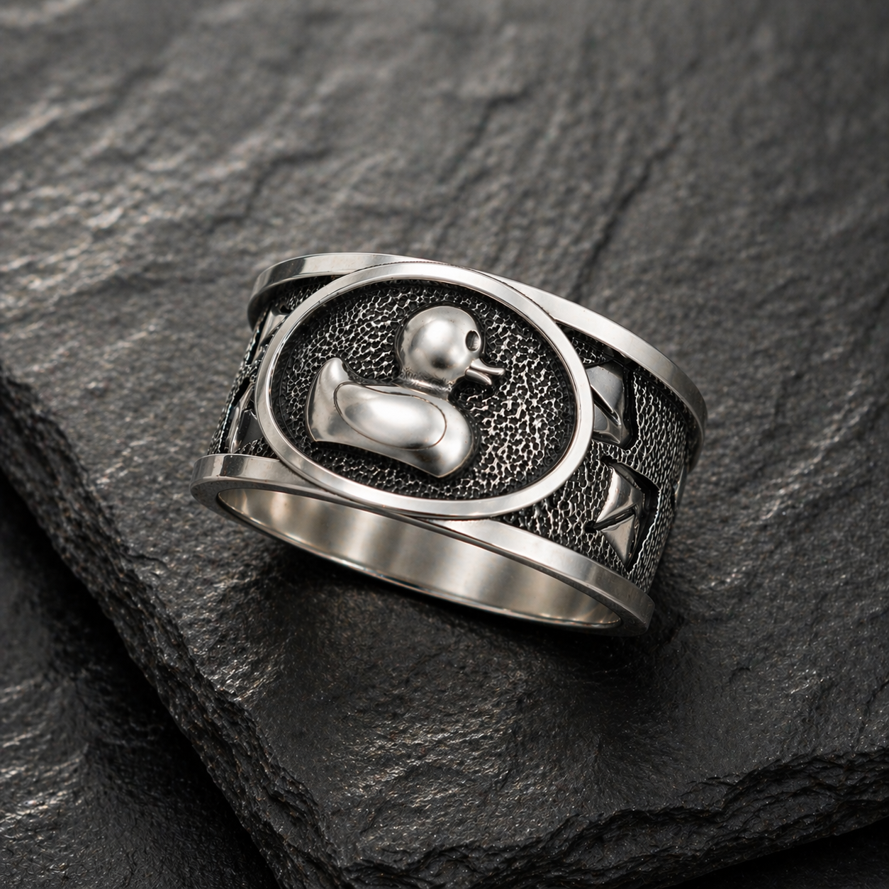 A silver ring with a metal duck design on a textured surface.