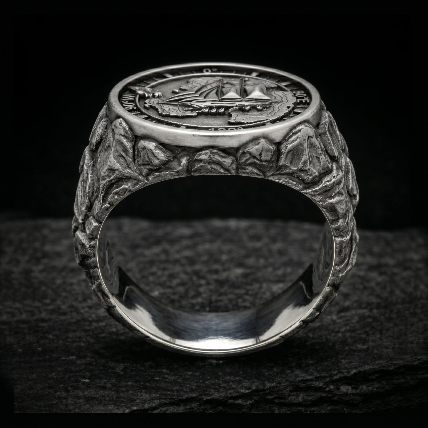 Antarctica explorer signet ring in solid sterling silver 925 featuring a detailed sailing ship over a map of Antarctica, surrounded by directional markings. The ring has a deep oxidized finish with strong contrast, highlighting fine handcrafted textures and relief details. Shot on a dark stone background to emphasize its bold, masculine design, adventure theme, and expedition spirit. Unique nautical jewelry piece inspired by travel, ocean journeys, and exploration beyond the known world.