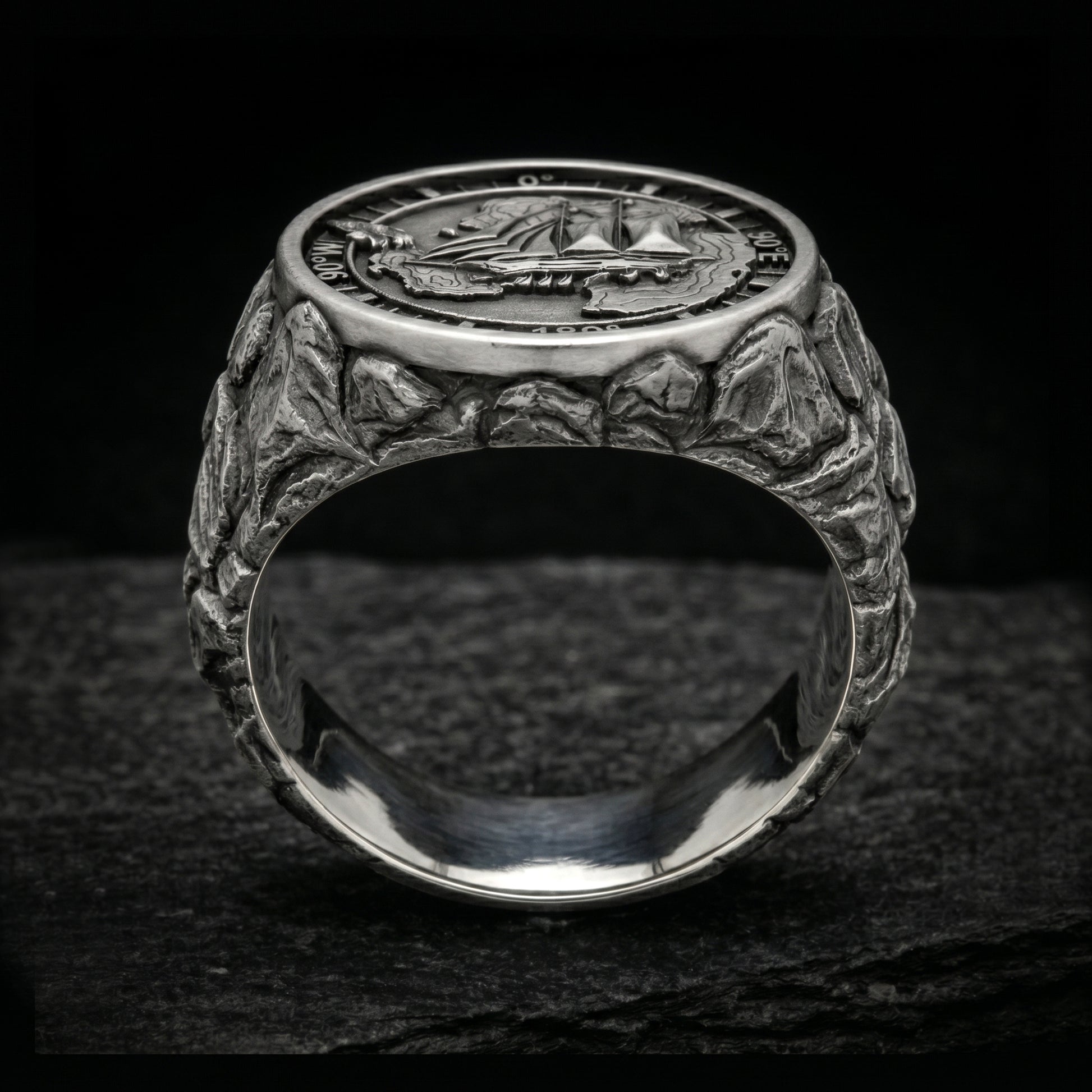 Antarctica explorer signet ring in solid sterling silver 925 featuring a detailed sailing ship over a map of Antarctica, surrounded by directional markings. The ring has a deep oxidized finish with strong contrast, highlighting fine handcrafted textures and relief details. Shot on a dark stone background to emphasize its bold, masculine design, adventure theme, and expedition spirit. Unique nautical jewelry piece inspired by travel, ocean journeys, and exploration beyond the known world.