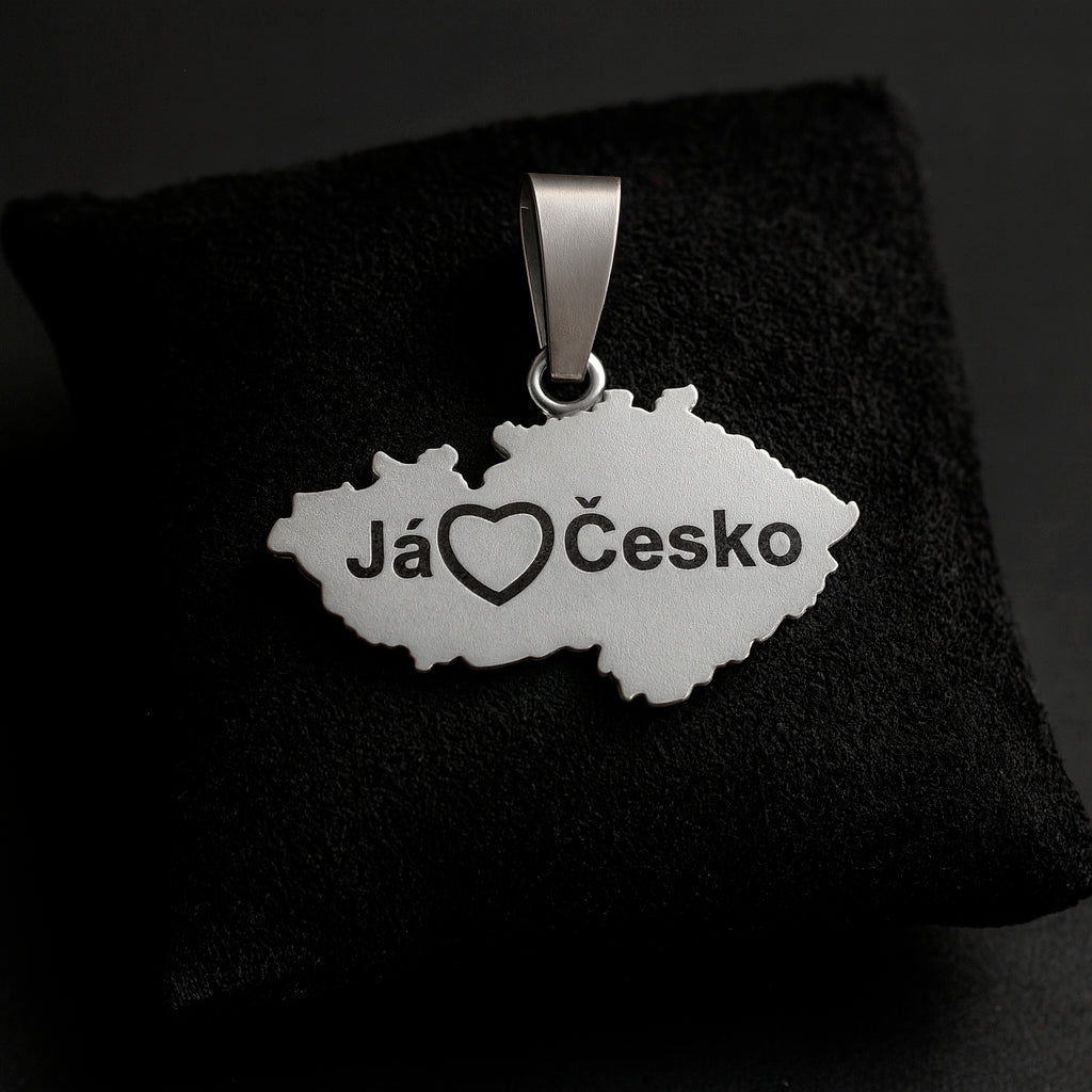 A handcrafted sterling silver pendant shaped like the map of the Czech Republic with the engraved phrase “Já❤️Česko.” The pendant measures 27 mm in width and 24 mm in height with the bail, weighs 3 g, and features a smooth polished edge with dark engraved details. It is displayed resting on a soft dark fabric cushion with gentle lighting that highlights the texture of the metal and the elegant contours of the design.