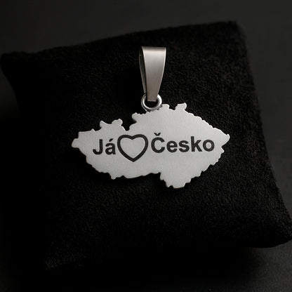 A handcrafted sterling silver pendant shaped like the map of the Czech Republic with the engraved phrase “Já❤️Česko.” The pendant measures 27 mm in width and 24 mm in height with the bail, weighs 3 g, and features a smooth polished edge with dark engraved details. It is displayed resting on a soft dark fabric cushion with gentle lighting that highlights the texture of the metal and the elegant contours of the design.