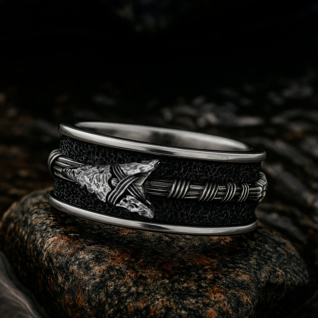 A silver ring with a black and silver design, featuring a pointed arrow on the band, is resting on a rock.