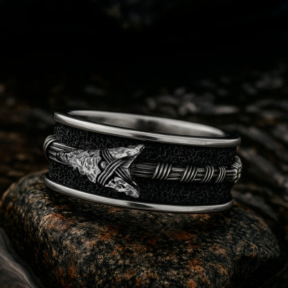 A silver ring with a black and silver design, featuring a pointed arrow on the band, is resting on a rock.