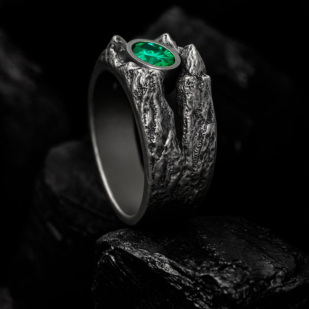 A silver ring with a large green gemstone, possibly an emerald, is displayed against a dark background.