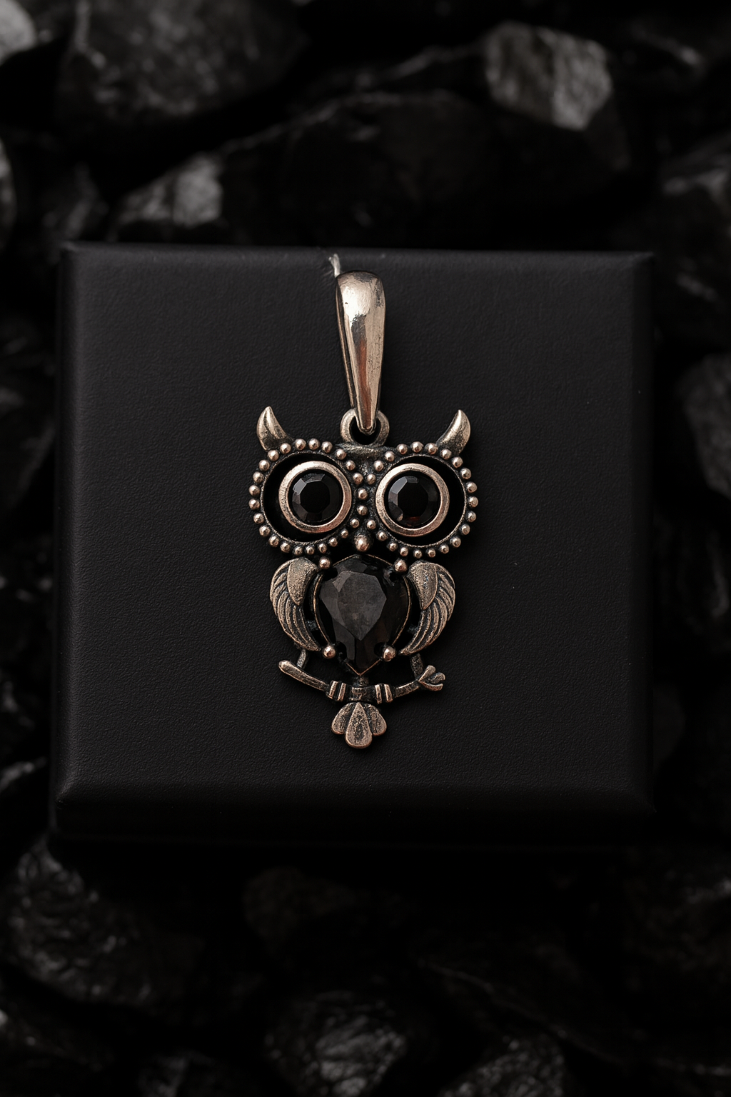 A silver owl pendant with black gemstones is displayed against a black background.