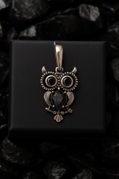 A silver owl pendant with black gemstones is displayed against a black background.