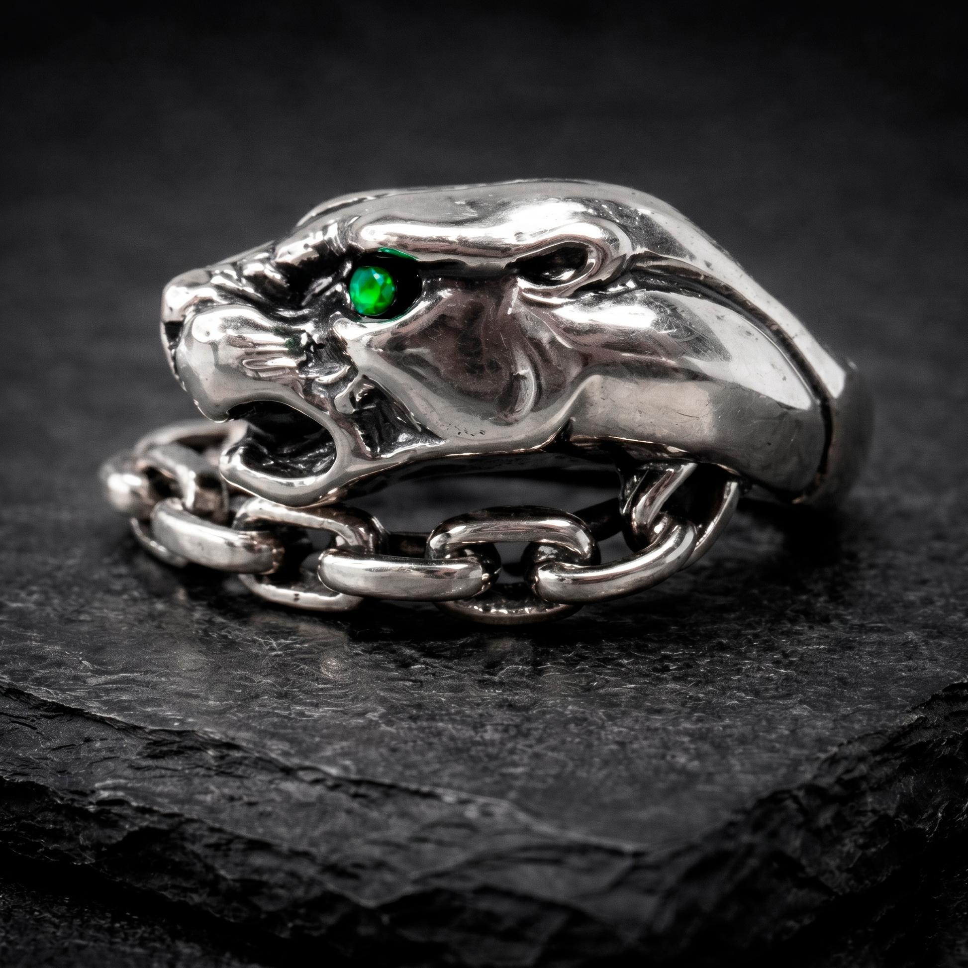 A silver ring with a green gemstone, resembling a panther's head, is displayed on a dark surface.