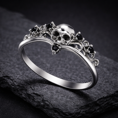 A silver ring with a skull design and black gemstones, placed on a dark surface.