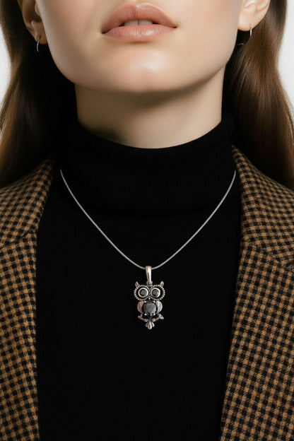 A woman wearing a black turtleneck sweater and a gold and black checkered jacket, with a silver pendant necklace featuring an owl pendant.