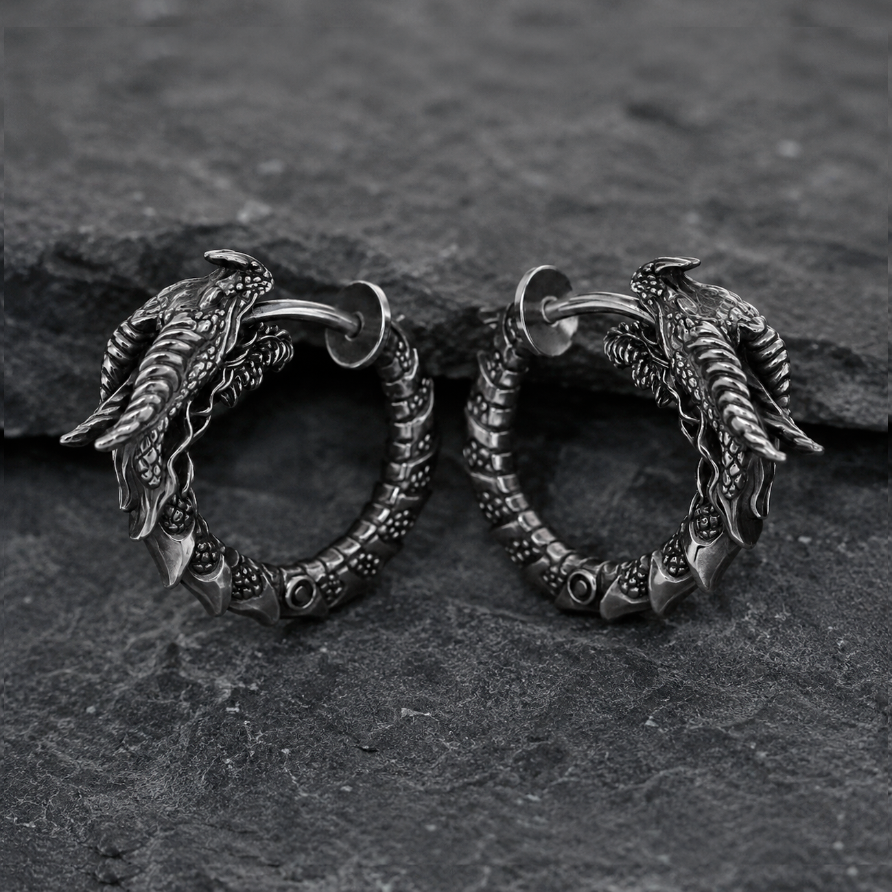 Pair of handcrafted dragon hoop earrings in oxidized sterling silver 925 by INDIGO Jewelry, photographed on a dark textured stone background. Detailed gothic earrings featuring sculpted dragon heads, textured scales, and segmented hoops with blackened silver finish for a vintage look. Fantasy-inspired silver jewelry with medieval and mythical aesthetics, designed for gothic style, dragon symbolism, and statement alternative fashion.