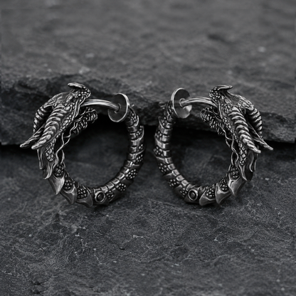 Pair of handcrafted dragon hoop earrings in oxidized sterling silver 925 by INDIGO Jewelry, photographed on a dark textured stone background. Detailed gothic earrings featuring sculpted dragon heads, textured scales, and segmented hoops with blackened silver finish for a vintage look. Fantasy-inspired silver jewelry with medieval and mythical aesthetics, designed for gothic style, dragon symbolism, and statement alternative fashion.