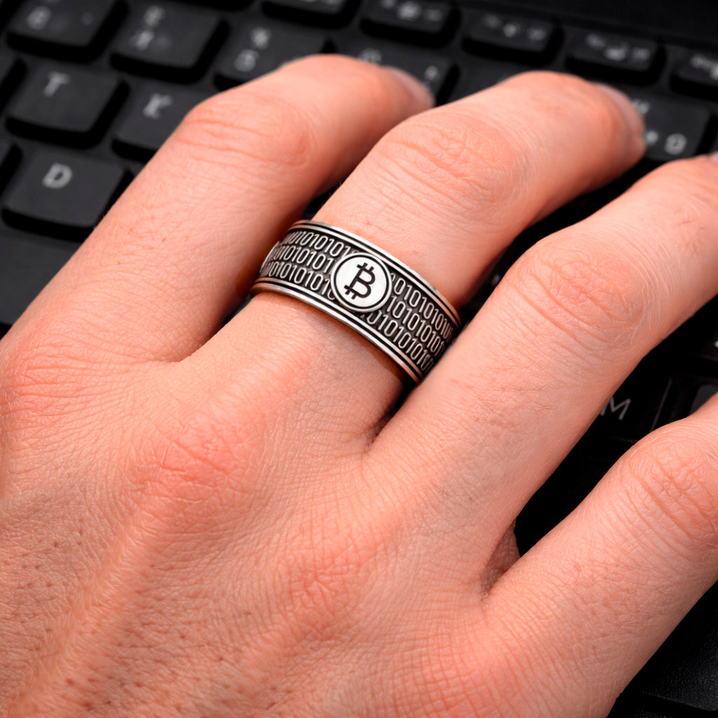 A close-up image of a person's hand wearing a ring with a Bitcoin symbol on it, resting on a computer keyboard. Sterling silver 925 bitcoin ring with blackened finish, wide band crypto ring featuring binary code and bitcoin symbol