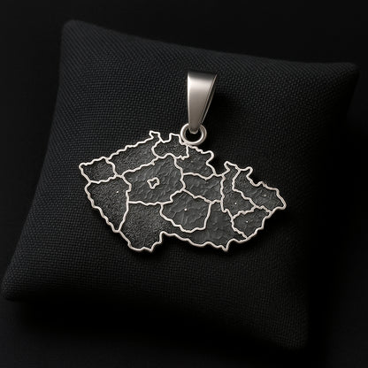 A handcrafted sterling silver pendant shaped like the map of the Czech Republic with the engraved phrase “Já❤️Česko.” The pendant measures 27 mm in width and 24 mm in height with the bail, weighs 3 g, and features a smooth polished edge with dark engraved details. It is displayed resting on a soft dark fabric cushion with gentle lighting that highlights the texture of the metal and the elegant contours of the design.