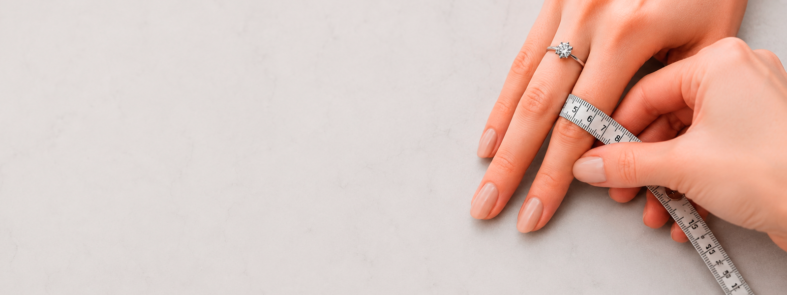 Woman measuring ring size at home using a measuring tape while wearing a solitaire engagement ring. Close-up ring size guide image showing how to measure finger size accurately for rings. Includes visual reference for ring size measurement, ring size chart and ring sizing at home for women and men by INDIGO Jewelry Atelier.